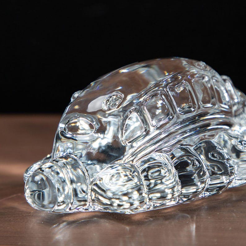 May include: A clear glass sculpture of a bus, showcasing intricate details. The bus features windows, a rounded front, and detailed wheel wells. The transparent material allows light to pass through, creating a shimmering effect. The sculpture is set against a dark background.