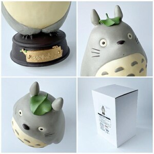 May include: A collection of Totoro figurines in various poses. The figurines are gray with cream-colored bellies and green leaf accents. One figurine is on a dark brown base with Japanese text. A white box with a Totoro illustration is also shown.
