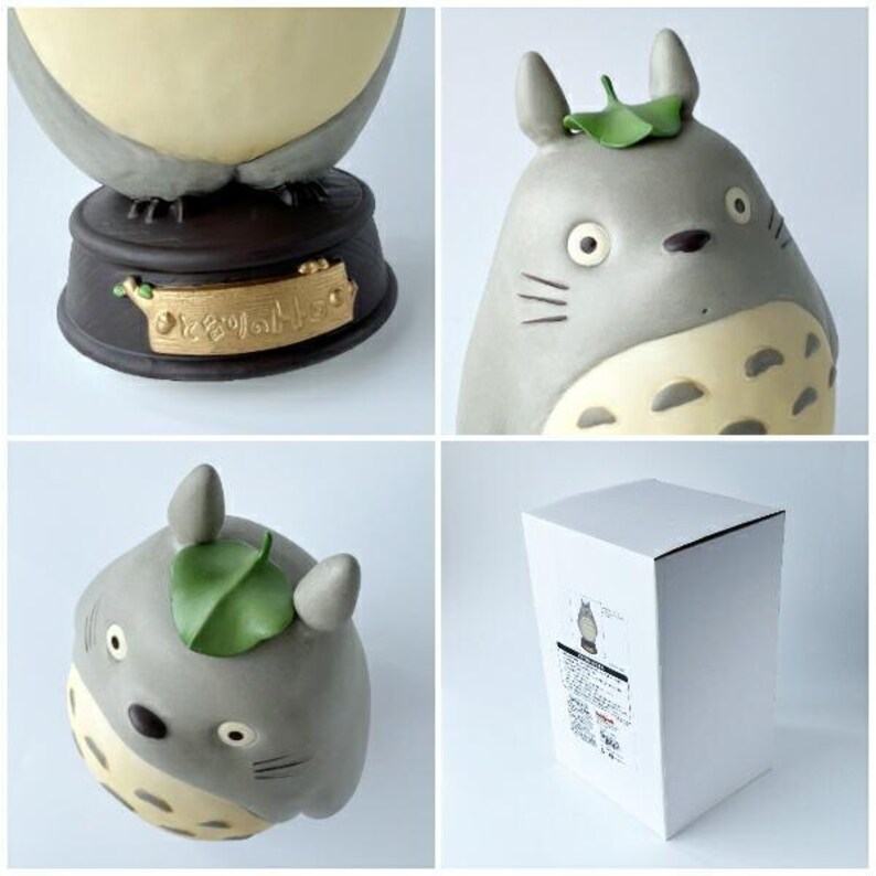 May include: A collection of Totoro figurines in various poses. The figurines are gray with cream-colored bellies and green leaf accents. One figurine is on a dark brown base with Japanese text. A white box with a Totoro illustration is also shown.