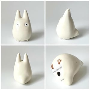 K&ouml;nnte beinhalten: Vier Bilder einer cremefarbenen Keramikfigur. Die Figur hat einen abgerundeten K&ouml;rper, zwei hohe Ohren und kleine Gesichtsz&uuml;ge. Auf der Unterseite der Figur steht "Studio Ghibli".