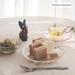 May include: A black cat figurine with a red bow, a clear glass teacup and saucer with a purple handle and rim, a clear glass bowl containing salad, and a clear glass plate holding four small sandwiches, with a metal fork in the foreground.