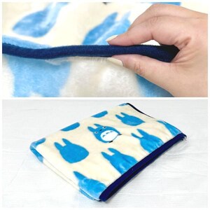 May include: A folded blanket featuring a pattern of blue cartoon characters on a cream background. The blanket has a dark blue border and is made of a soft, plush material. The image shows a close-up of the blanket's edge.
