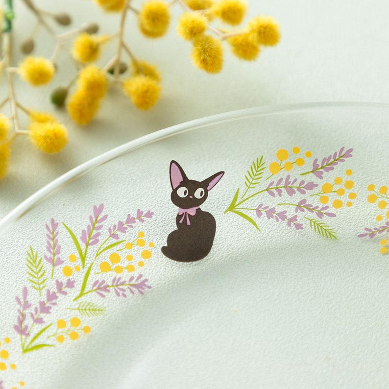 May include: A clear glass plate featuring a central illustration of a small black cat with a pink bow around its neck, surrounded by a decorative pattern of yellow mimosa flowers and light purple sprigs of lavender arranged in a curved arc.