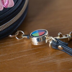 May include: A close-up of a black zippered pouch with a silver-coloured retractable key reel. The reel has a colourful design with blue, green, red, and yellow segments. A black strap and gold-coloured clasp are also visible.
