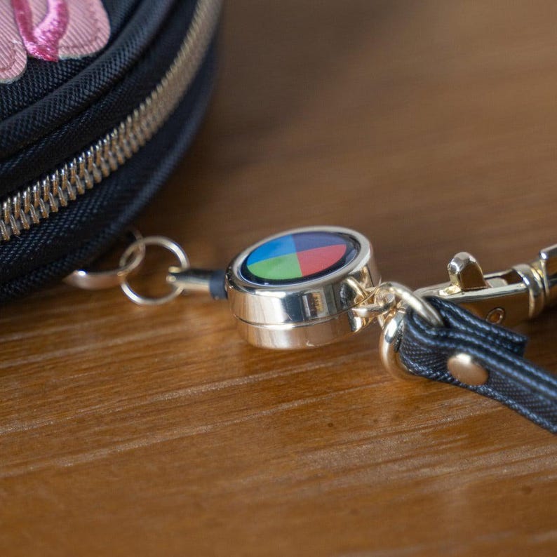 May include: A close-up of a black zippered pouch with a silver-coloured retractable key reel. The reel has a colourful design with blue, green, red, and yellow segments. A black strap and gold-coloured clasp are also visible.