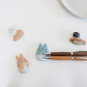 Puede incluir: Cuatro reposaplumas de cer&aacute;mica con forma de personajes de dibujos animados, incluyendo dos figuras color canela con orejas de conejo, una figura azul con orejas puntiagudas y una figura de cabeza oscura con texto japon&eacute;s, junto a dos bol&iacute;grafos de madera.