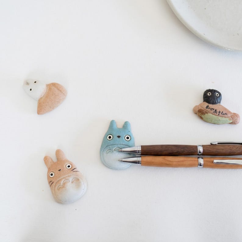 Puede incluir: Cuatro reposaplumas de cer&aacute;mica con forma de personajes de dibujos animados, incluyendo dos figuras color canela con orejas de conejo, una figura azul con orejas puntiagudas y una figura de cabeza oscura con texto japon&eacute;s, junto a dos bol&iacute;grafos de madera.