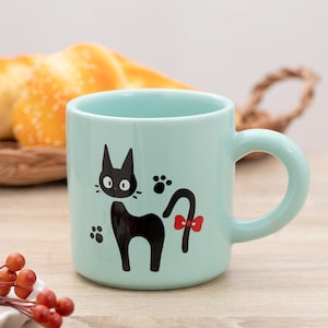 May include: A light mint green ceramic mug featuring a black cat illustration with large white eyes, whiskers, and a red bow on its tail. The design includes two black paw prints on either side of the cat. The mug has a smooth, rounded handle.