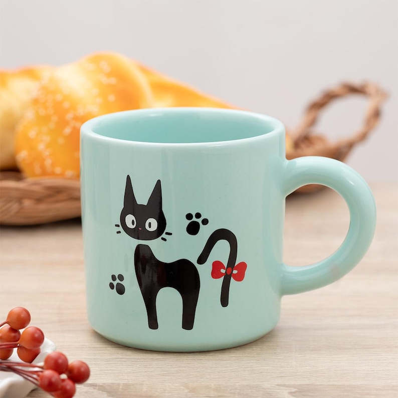 May include: A light mint green ceramic mug featuring a black cat illustration with large white eyes, whiskers, and a red bow on its tail. The design includes two black paw prints on either side of the cat. The mug has a smooth, rounded handle.