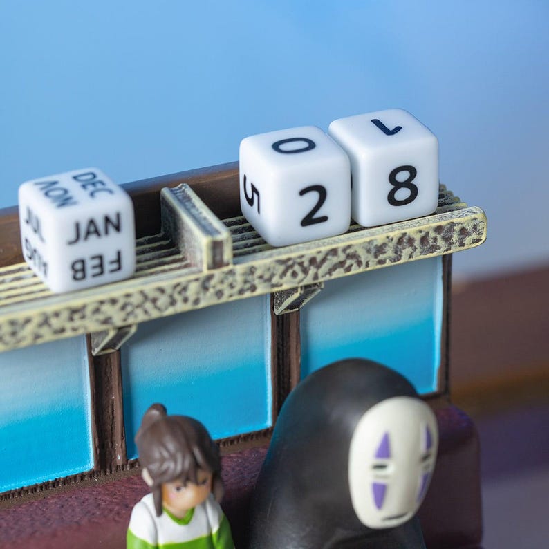 May include: A miniature decorative calendar featuring a scene with figurines. The calendar displays the date 05/28. The figurines include a girl and a character with a white mask. The calendar has months printed on the side.