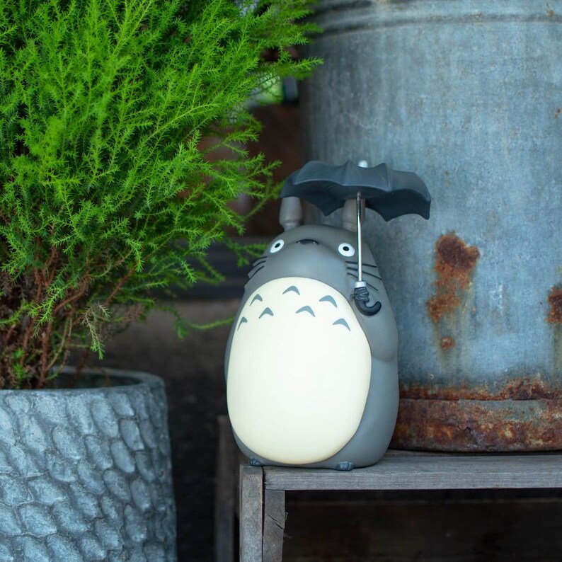 Puede incluir: Una figura de Totoro gris con una barriga de color crema que presenta marcas de chevr&oacute;n en gris oscuro. El personaje tiene dos orejas puntiagudas, ojos blancos grandes con pupilas negras y sostiene un peque&ntilde;o paraguas negro con mango plateado.