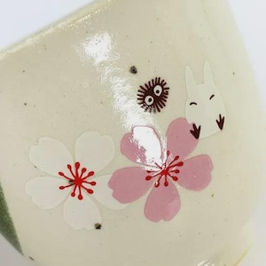 May include: Cream-colored ceramic cup with a floral design. The cup features white and pink cherry blossoms, a cartoon rabbit, and a small, round, dark brown creature. The cup has a slightly textured surface.