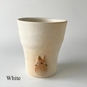May include: A white ceramic mug with a textured surface and a Totoro character design. The mug has a curved shape and a feather-like etched design. The word "White" is printed at the bottom of the mug.