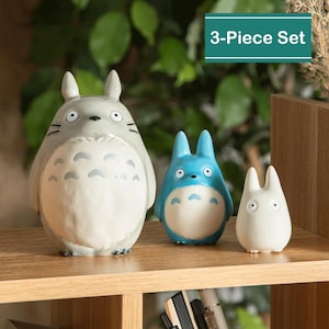 May include: A 3-Piece Set of Totoro figurines in gray, blue, and white. The largest figurine is gray with a white belly and black markings. The middle figurine is blue with a white belly. The smallest figurine is white. The set is displayed on a wooden shelf.