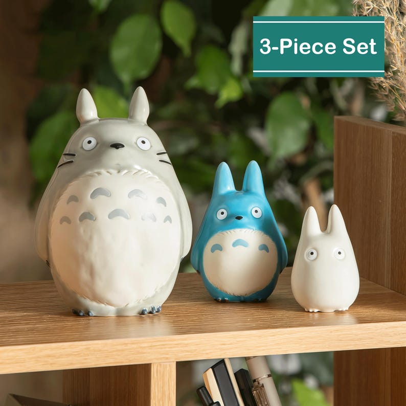 May include: A 3-Piece Set of Totoro figurines in gray, blue, and white. The largest figurine is gray with a white belly and black markings. The middle figurine is blue with a white belly. The smallest figurine is white. The set is displayed on a wooden shelf.