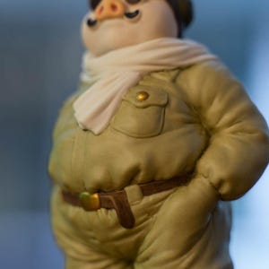 May include: A ceramic pig figurine wearing a light green military-style jacket with a brown belt and a white scarf. The pig has a black moustache and a small hat. The jacket has a pocket with a gold button.
