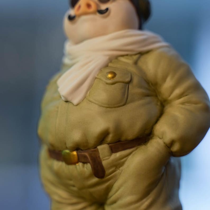 May include: A ceramic pig figurine wearing a light green military-style jacket with a brown belt and a white scarf. The pig has a black moustache and a small hat. The jacket has a pocket with a gold button.