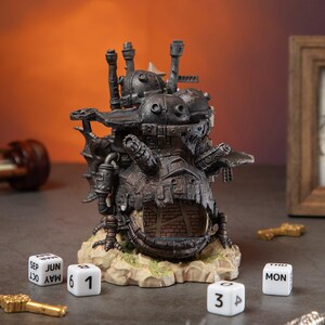 May include: A detailed, dark gray miniature of a fantastical, steampunk-style building with chimneys and mechanical details. The base is a light tan with green accents. White dice with numbers and months are scattered around the base.