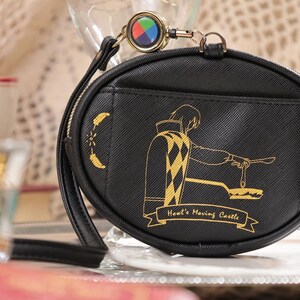 May include: Black, oval coin purse with a gold zip and wrist strap. The purse features a gold illustration of a character from "Howl's Moving Castle" and the text "Howl's Moving Castle". A colourful charm is attached.