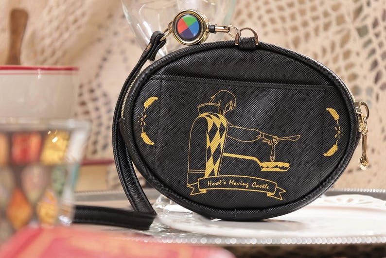 May include: Black, oval coin purse with a gold zip and wrist strap. The purse features a gold illustration of a character from "Howl's Moving Castle" and the text "Howl's Moving Castle". A colourful charm is attached.