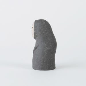 Puede incluir: Una peque&ntilde;a figura de color gris oscuro con textura y un rostro ovalado, p&aacute;lido y liso. La figura presenta una silueta redondeada y encapuchada con un dise&ntilde;o sencillo, que incluye dos peque&ntilde;os puntos negros como ojos y una marca violeta.