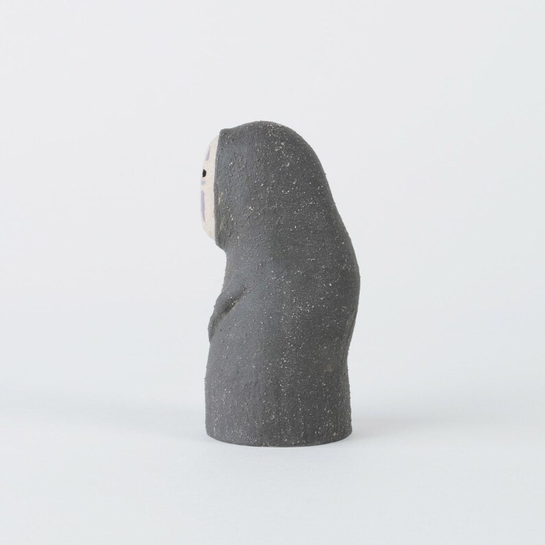 Puede incluir: Una peque&ntilde;a figura de color gris oscuro con textura y un rostro ovalado, p&aacute;lido y liso. La figura presenta una silueta redondeada y encapuchada con un dise&ntilde;o sencillo, que incluye dos peque&ntilde;os puntos negros como ojos y una marca violeta.