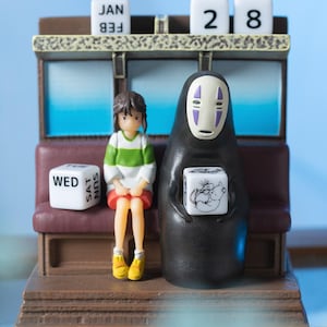 May include: A decorative figurine featuring characters from a popular animated film. The figurine includes a girl in a green and white striped shirt and a black figure with a white mask. A calendar displays "JAN 28" and "WED" and "SUN SAT".
