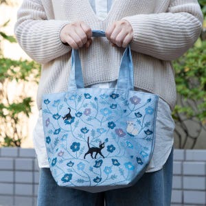 May include: A light blue tote bag with embroidered floral designs and black cats. The bag has a light blue canvas fabric with a white cotton lining.