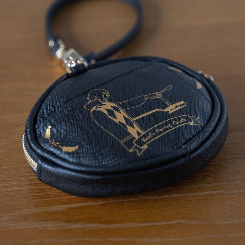 May include: A navy blue round coin purse with a gold zip and wrist strap. The purse features a gold illustration of a character and the text "Howl's Moving Castle". The design includes gold crescent moons.
