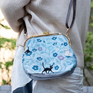 May include: A light blue handbag with a floral pattern and black cat embroidery. The bag has a gold-colored clasp and a brown shoulder strap. The floral design includes blue and purple flowers.