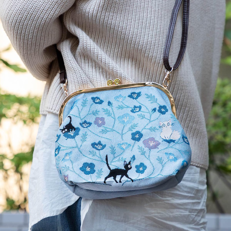 May include: A light blue handbag with a floral pattern and black cat embroidery. The bag has a gold-colored clasp and a brown shoulder strap. The floral design includes blue and purple flowers.