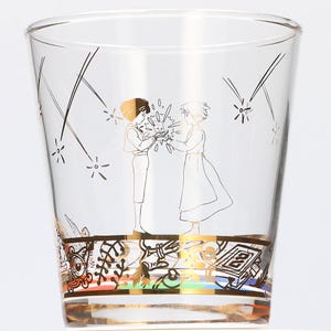 May include: Clear glass tumbler with a gold illustration of two figures and a small creature. The design features gold accents and a decorative band at the base with colorful details. The glass is ideal for serving beverages.