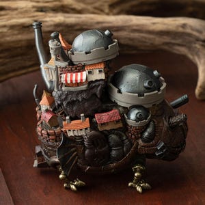 May include: A detailed, fantasy-themed figurine depicting a mechanical creature with a town built on its back. The figurine features a brown and bronze color scheme, with miniature buildings, turrets, and a chimney. The creature has clawed feet and a wing.