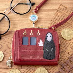 Pode incluir: Uma carteira bordô com fecho de zíper, com um design de vagão de trem com um personagem Sem Rosto e uma criatura branca. Inclui uma alça combinando e um pingente. A carteira está em uma superfície de madeira.