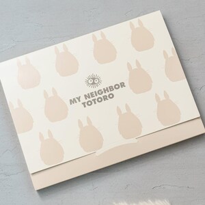 May include: A light beige box featuring a pattern of light brown Totoro silhouettes. The text "MY NEIGHBOR TOTORO" is printed in gray. The box has a light brown base.