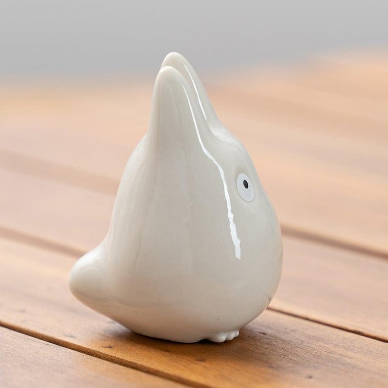K&ouml;nnte beinhalten: Eine wei&szlig;e Keramikfigur einer stilisierten Kreatur mit rundem K&ouml;rper und spitzem Kopf. Die Figur hat ein einzelnes Auge mit schwarzer Pupille und steht auf einer Holzoberfl&auml;che.
