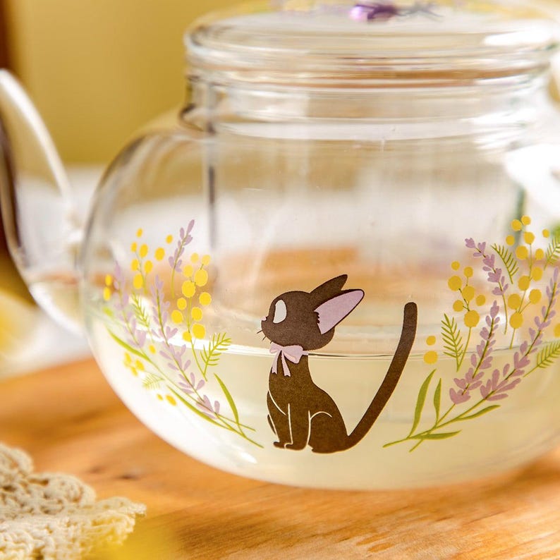 Puede incluir: Una tetera de cristal transparente con el gr&aacute;fico de un gato marr&oacute;n con un lazo rosa, rodeado de ilustraciones de flores de mimosa amarillas y ramas de lavanda moradas. La tetera incluye una tapa de cristal a juego con un peque&ntilde;o detalle floral.