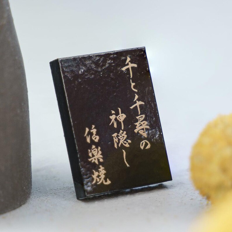 Puede incluir: Una placa rectangular negra con un acabado texturizado y brillante. La superficie presenta caracteres de caligraf&iacute;a japonesa de color dorado escritos verticalmente. La placa est&aacute; colocada en posici&oacute;n vertical, mostrando el texto claramente sobre el fondo oscuro.