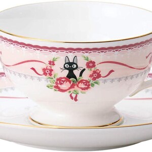 May include: A white teacup and saucer set with a pink floral design and gold trim. The teacup features a black cat illustration surrounded by roses. The saucer and cup have a decorative pink ribbon and lace design.