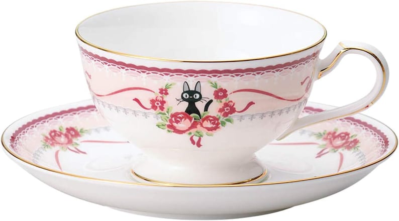 May include: A white teacup and saucer set with a pink floral design and gold trim. The teacup features a black cat illustration surrounded by roses. The saucer and cup have a decorative pink ribbon and lace design.