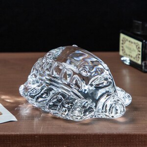 May include: A clear glass turtle figurine with detailed shell and limbs. The figurine has a textured surface and is sitting on a brown surface. The Studio Ghibli logo is visible on the figurine.