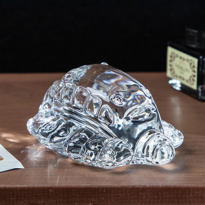 May include: A clear glass turtle figurine with detailed shell and limbs. The figurine has a textured surface and is sitting on a brown surface. The Studio Ghibli logo is visible on the figurine.