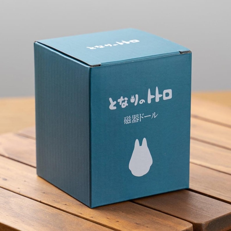 May include: A teal-colored box with Japanese text and a white silhouette of a character. The box is sitting on a wooden surface. The text on the box reads "となりのトトロ" and "磁器ドール".