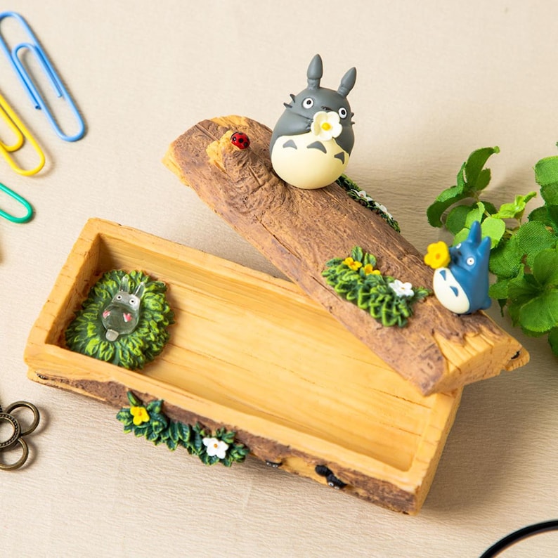 May include: A wooden box with a lid, decorated with miniature Totoro figures and floral accents. The lid is a piece of wood with a Totoro figure holding a flower. The box has a green creature figure inside.