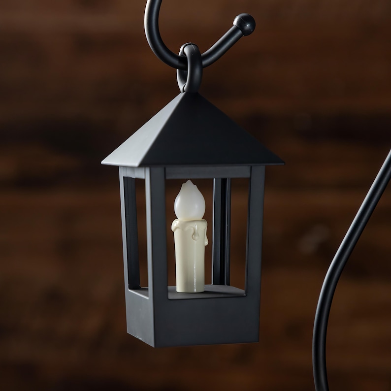 May include: A black metal lantern with a square base and a peaked roof. Inside the lantern is a cream-colored candle with a dripping wax design. The lantern is suspended from a curved black metal hook.