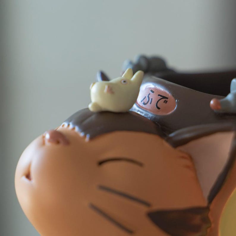 May include: Close-up of a tan and brown figurine of a cat-like character with a small, light yellow bunny on its head. The figurine has a serene expression and Japanese text on its collar.