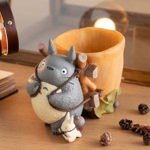 Puede incluir: Una figura gris de Totoro sostiene una maceta beige con una cuerda marrón. Una pequeña figura de conejo blanco está en la base. La maceta es beige con un diseño de tronco de árbol marrón. La escena está ambientada sobre una superficie de madera.