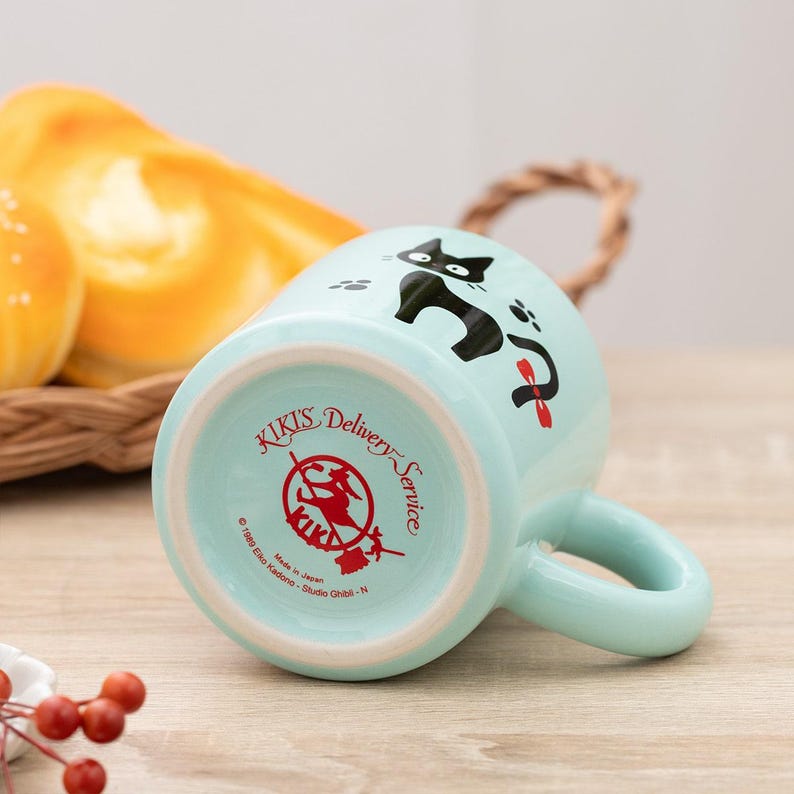 May include: A light blue ceramic mug featuring a black cat illustration with a red bow. The bottom of the mug displays the red text Kiki's Delivery Service, a circular logo of a witch on a broomstick, and the copyright 1989 Eiko Kadono - Studio Ghibli - N.