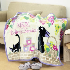 May include: A cream blanket with the text "Kiki's Delivery Service" and illustrations of a black cat, grapes, and a basket of flowers. The blanket has a purple border and is on a beige sofa.