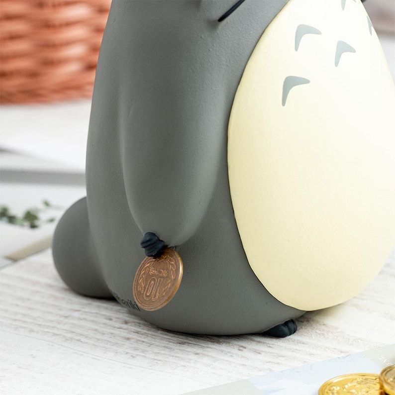 Puede incluir: Una hucha de cer&aacute;mica gris con forma de criatura y un gran vientre color crema con marcas de chevr&oacute;n gris oscuro. La figura tiene un cuerpo redondeado y sostiene una peque&ntilde;a moneda redonda de color cobre entre los dedos cerca de la base.
